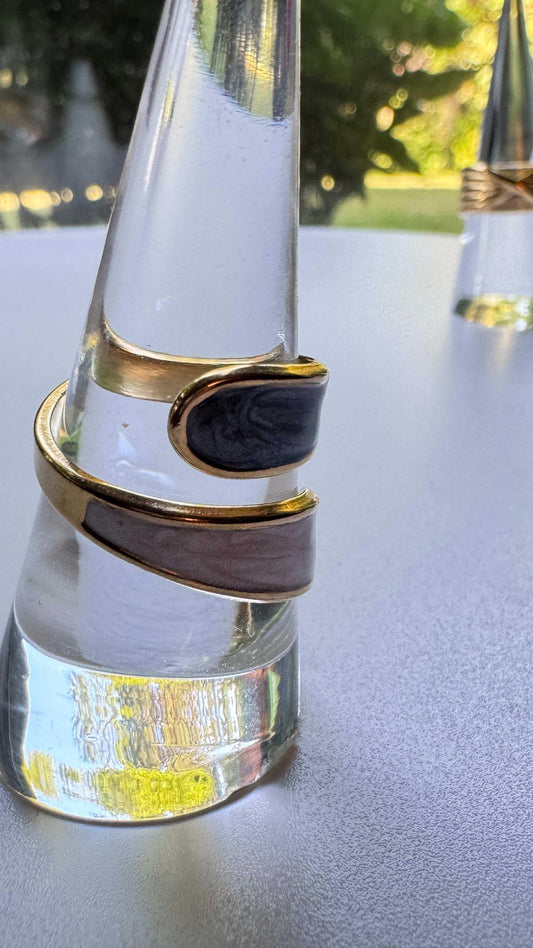 Gold adjustable ring with black and gray enamel on a clear display stand outdoors