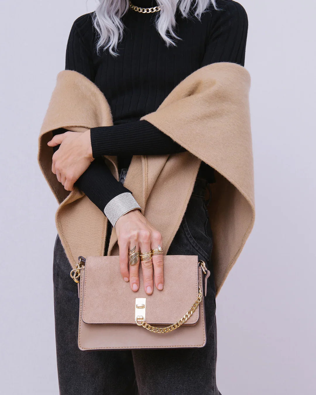 Woman holding beige suede crossbody bag with gold chain, wearing black sweater and camel shawl