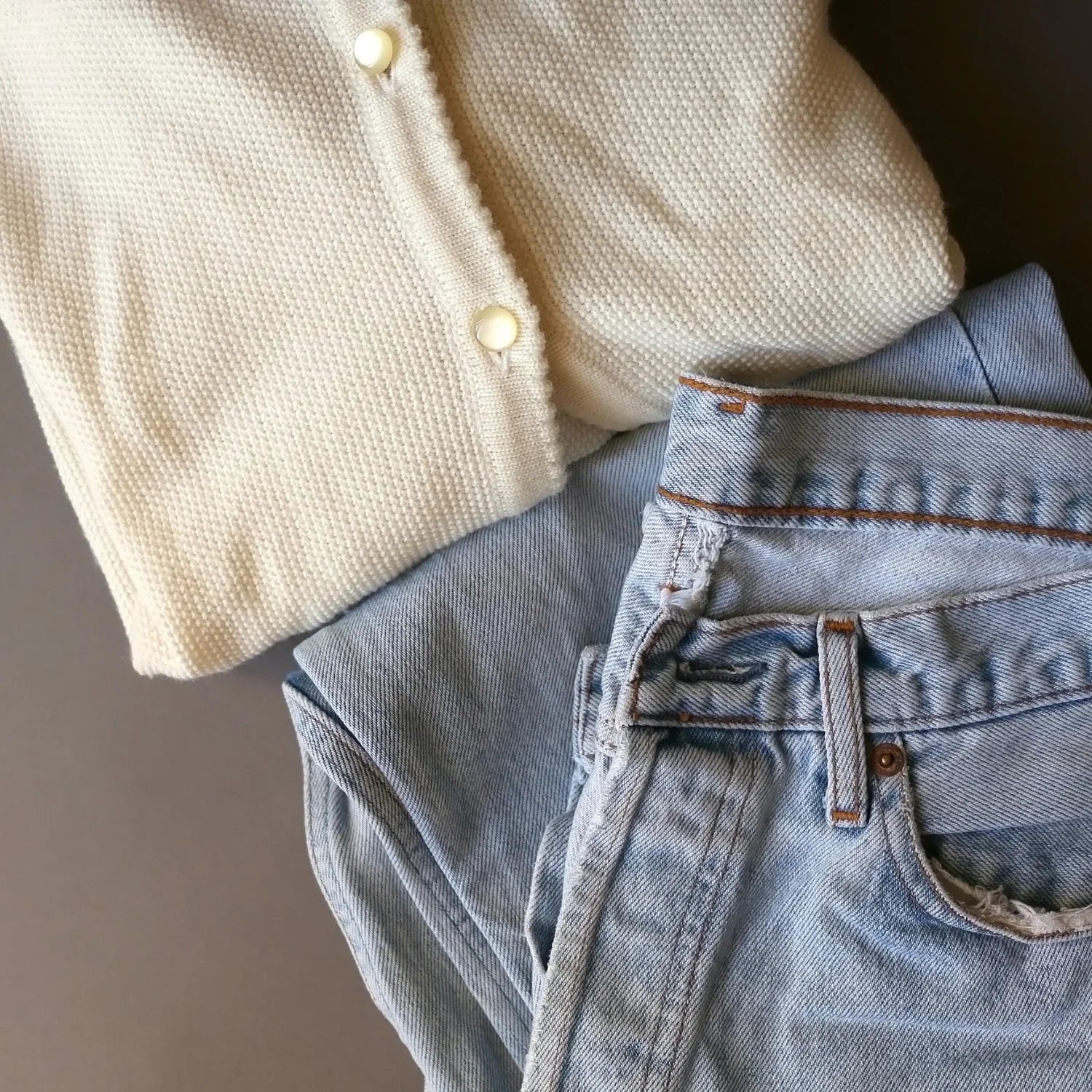 Folded cream knit sweater with buttons and light blue denim jeans on a neutral background
