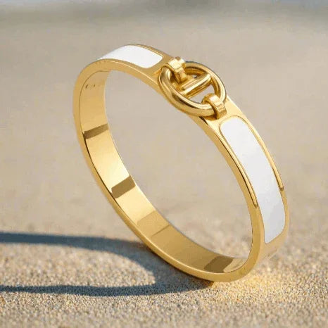 Gold clasp bracelet with white enamel inlay resting on sandy surface