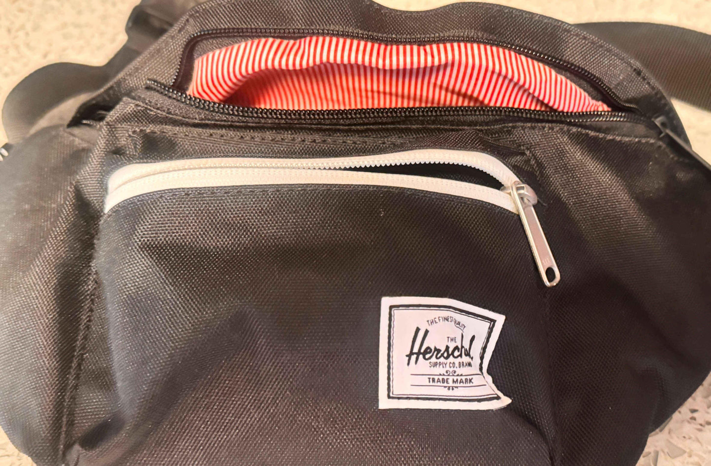 Black Herschel fanny pack with white zipper and red-striped lining on a speckled surface