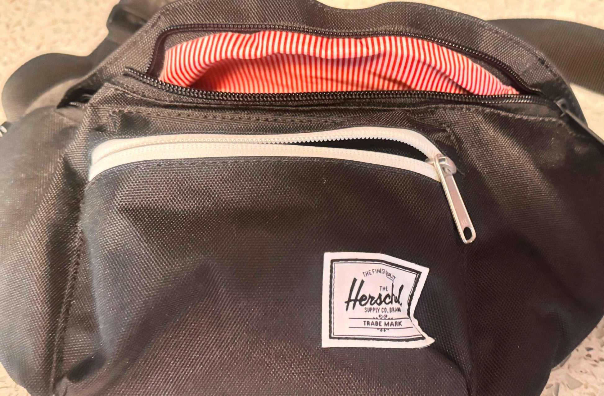 Black Herschel fanny pack with white zipper and red-striped lining on a speckled surface