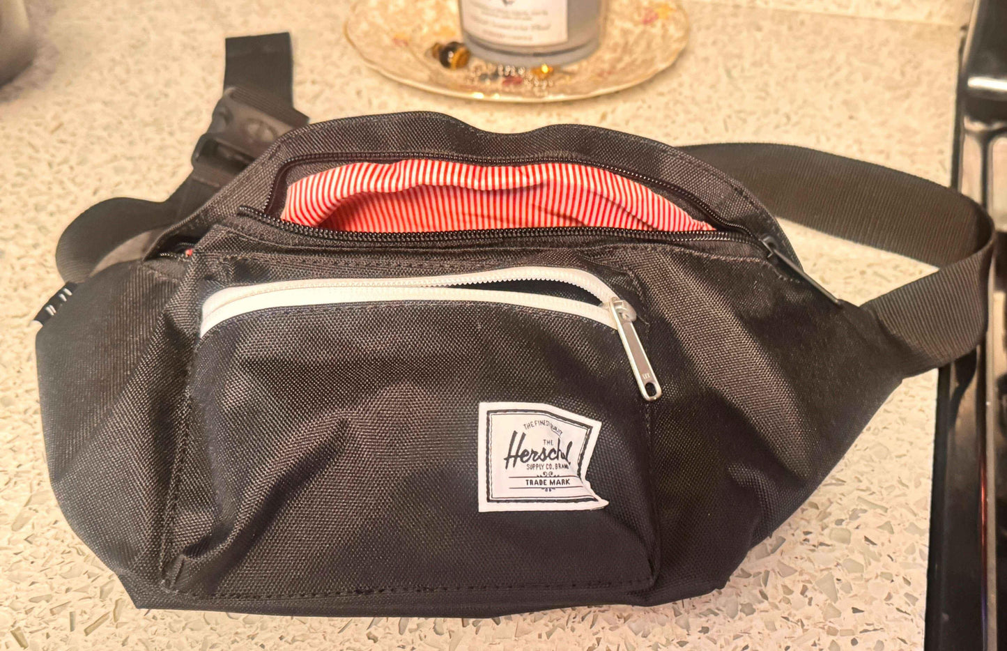 Black Herschel fanny pack with white zipper and red striped lining on a countertop.