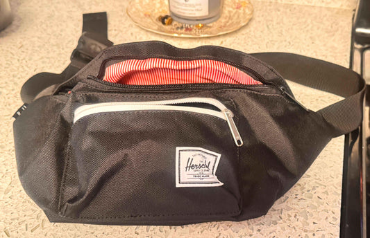 Black Herschel fanny pack with white zipper and red striped lining on a countertop.