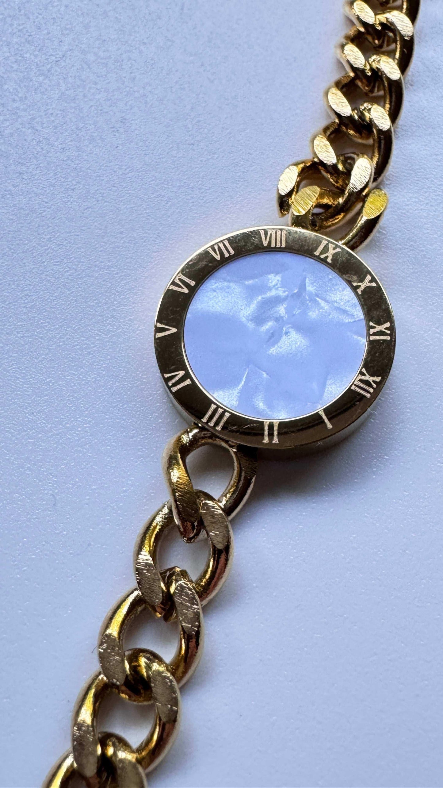 Gold chain bracelet with round Roman numeral bezel and light blue stone center on white surface